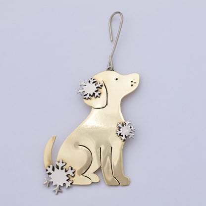 A gold-colored metal Christmas ornament shaped like a sitting dog, decorated with three silver-colored laser-cut snowflakes on its ears, chest, and tail, hanging from a silver wire hook against a light lavender background.