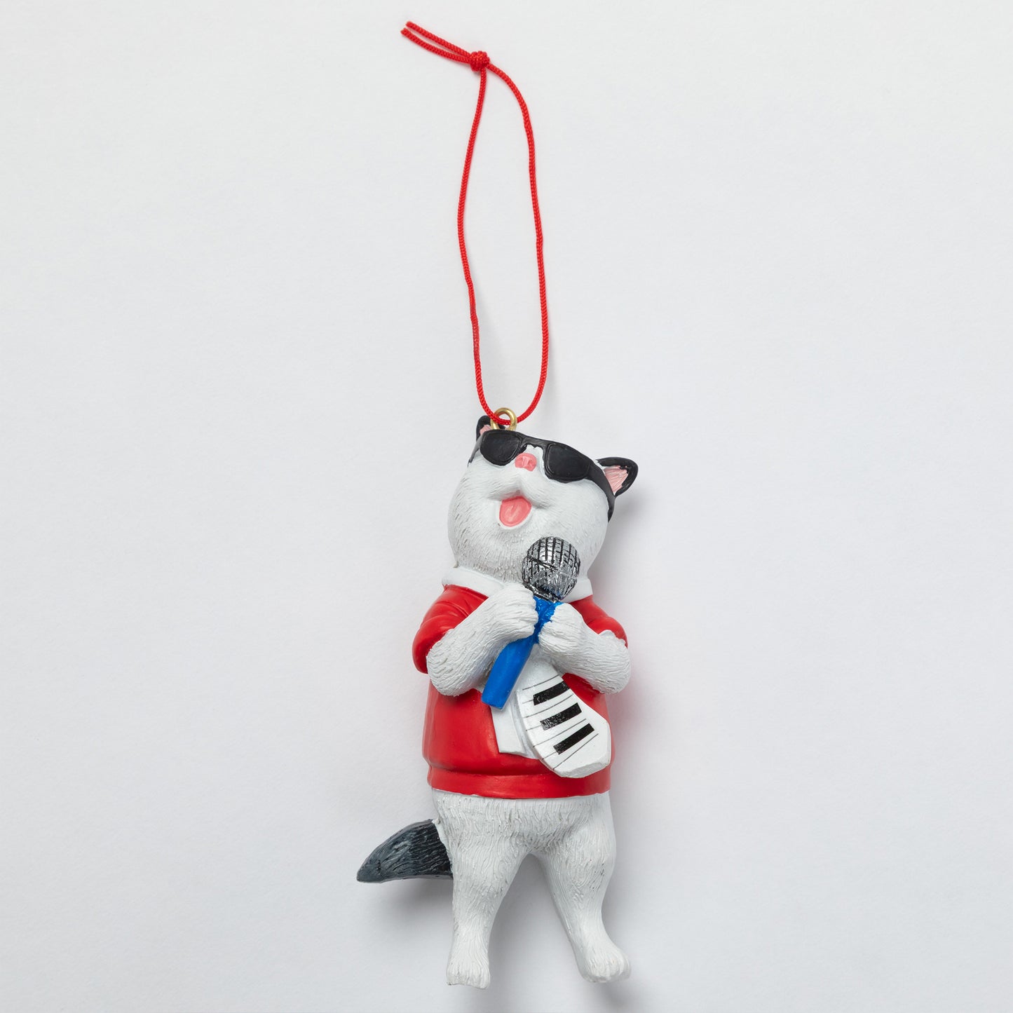 A small cat-shaped hanging ornament or figurine wearing a red shirt, blue tie, and white-and-black striped guitar, with a red hanging string at the top, photographed against a plain light background.