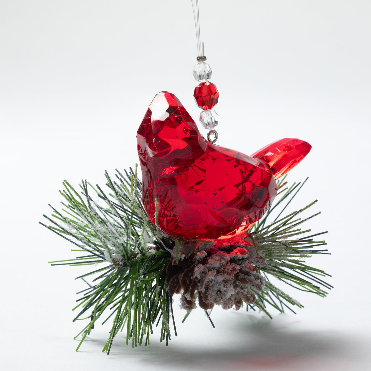 A red faceted bird Christmas ornament with a hanging loop, nestled among green pine needles and a small frosted brown pinecone on a white background.