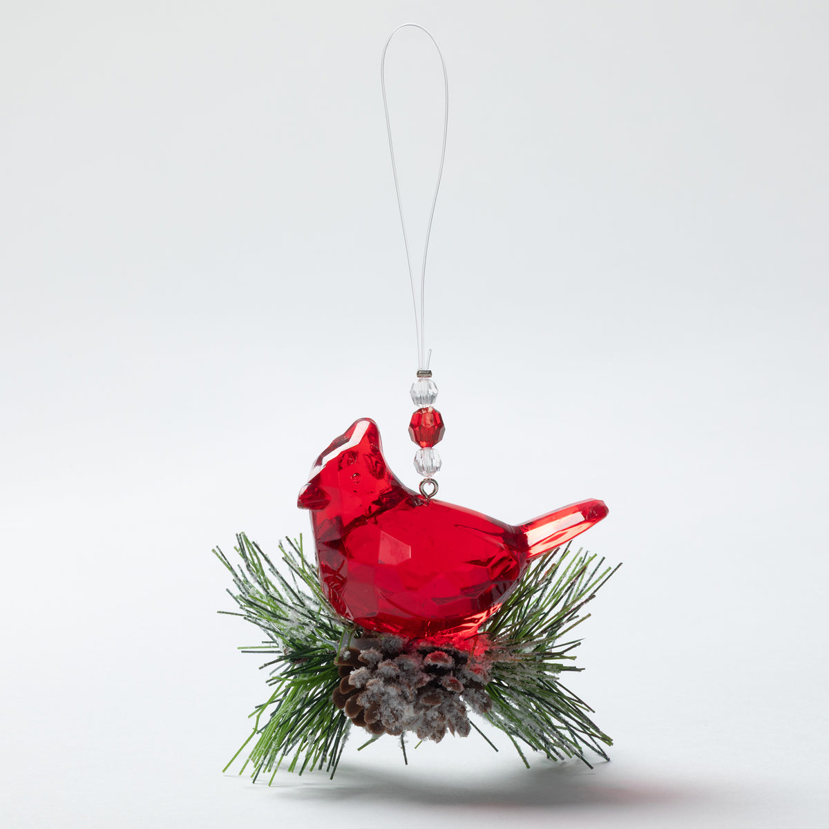 A red plastic cardinal Christmas ornament perched on green pine branches with a small pinecone, hanging from a clear string against a light gray background.