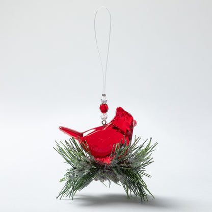 A red glass cardinal Christmas ornament perched on a green pine branch with a white hanging loop, shown against a light gray studio background.