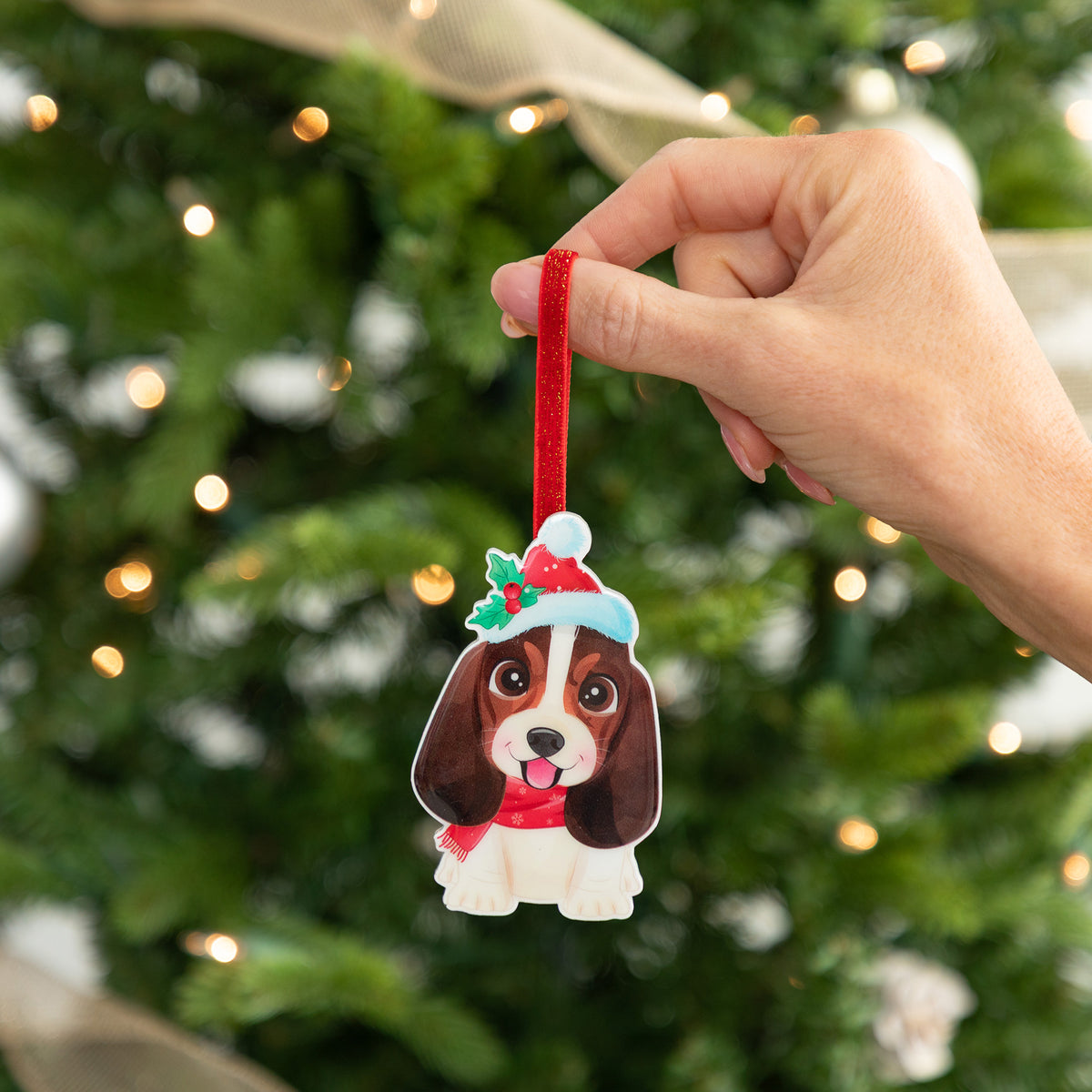 A hand holds a custom dog Christmas ornament shaped like a brown and white Cocker Spaniel wearing a red Santa hat and scarf, set against a blurred Christmas tree background with warm lights.