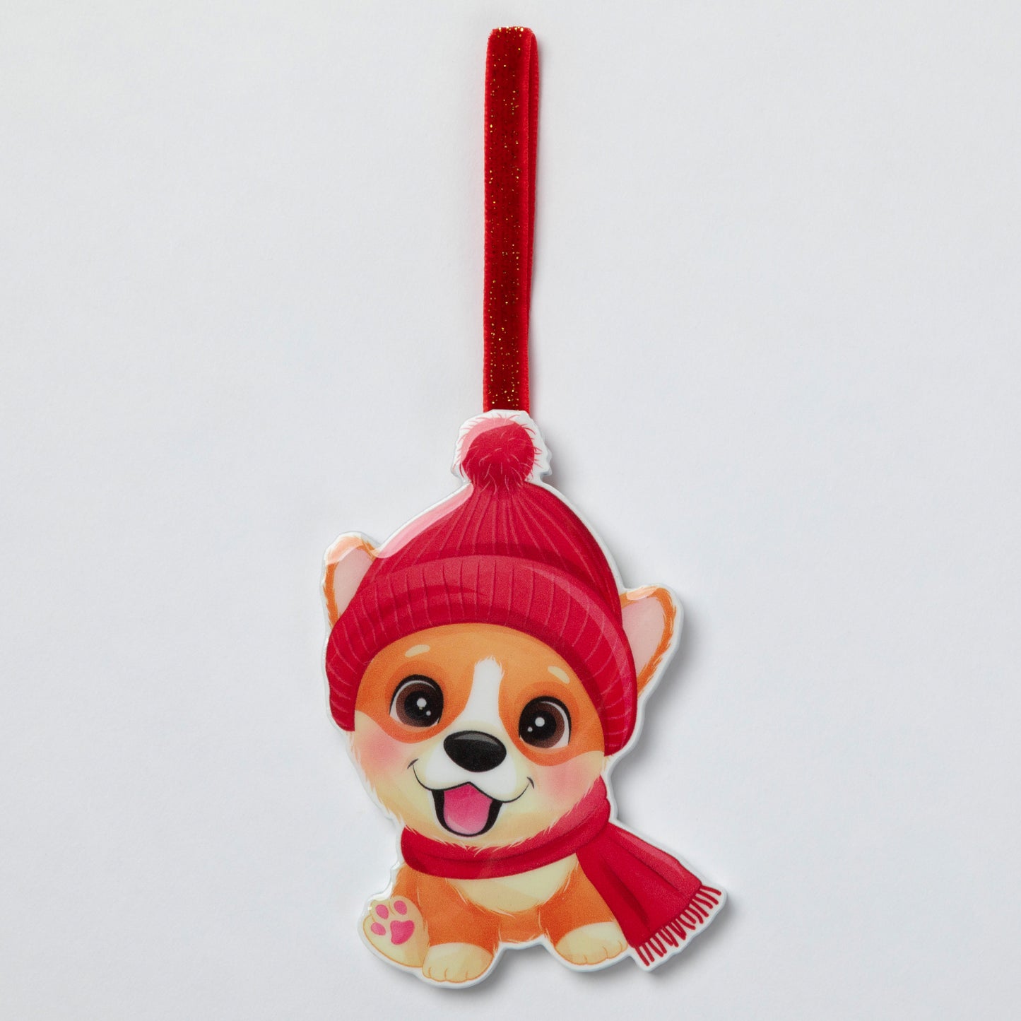 A cute cartoon dog hanging ornament wearing a red knit hat and matching red scarf, suspended from a red glittery ribbon on a light gray background.