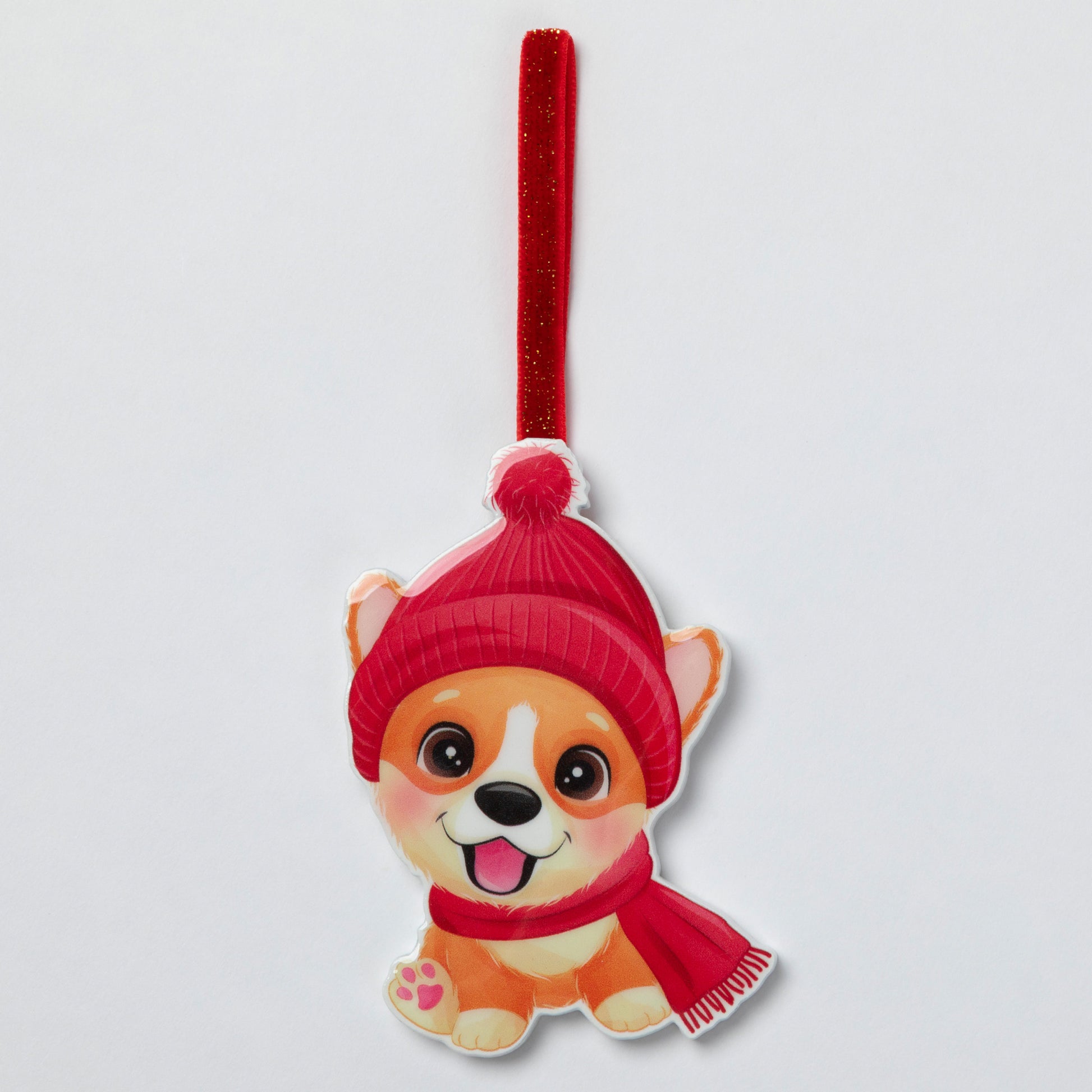 A cute cartoon dog hanging ornament wearing a red knit hat and matching red scarf, suspended from a red glittery ribbon on a light gray background.