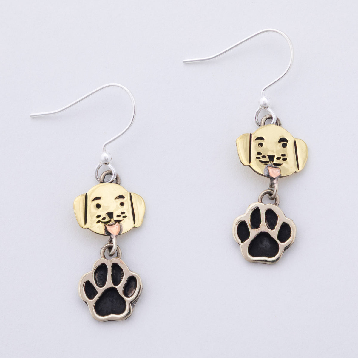 A pair of dog-themed dangle earrings featuring brass and silver dog head charms with black paw prints hanging below, shown on a plain light gray background.