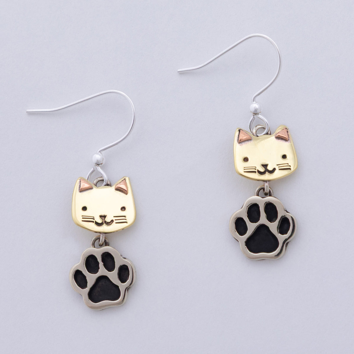 A pair of dangle earrings featuring a brass cat face charm and a silver paw print charm with black paw pads, shown on a light gray background.