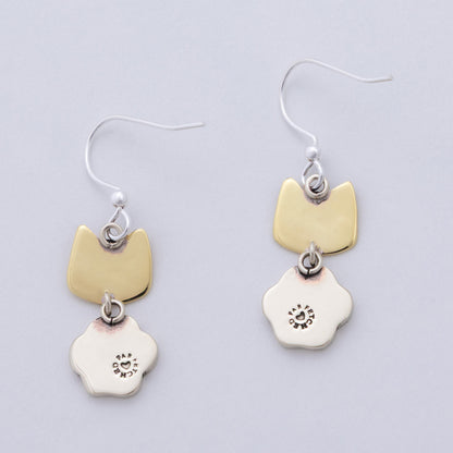 The back of a pair of brass and silver earrings, shaped like cat heads and paws.