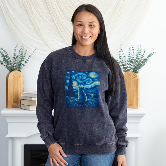 A woman wearing a dark gray acid-wash crewneck sweatshirt with a 'Starry Night' cat graphic on the front, standing in front of a white fireplace mantel decorated with plants.
