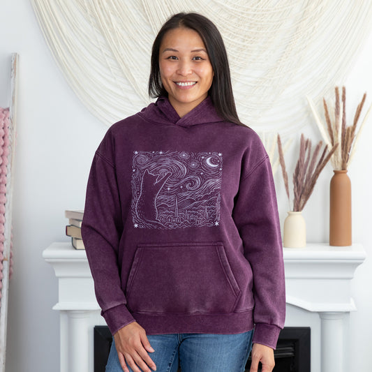 A woman wearing a purple hooded sweatshirt with a white line art graphic of a cat under a crescent moon, standing in front of a white fireplace mantel in a decorated room.