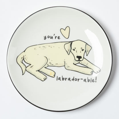 A round white plate with a black rim, featuring a hand-drawn illustration of a light cream Labrador puppy lying down with the text 'you're labrador-able!' and a small heart above it.
