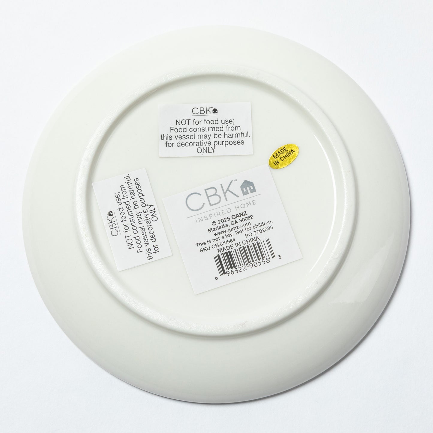 The underside of a white ceramic plate or shallow bowl, showing printed labels and a barcode sticker. The brand name CBK is clearly visible on the labels.