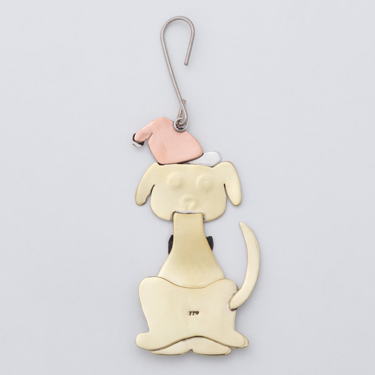 A handmade dog-shaped pendant made of three metals: a large gold-toned body, a rose gold hat, and a small white accent, hanging from a silver hook on a light gray background.