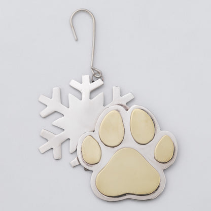 A silver metal ornament featuring a paw print with gold-colored pads and a separate snowflake cutout behind it, hanging against a light gray background.