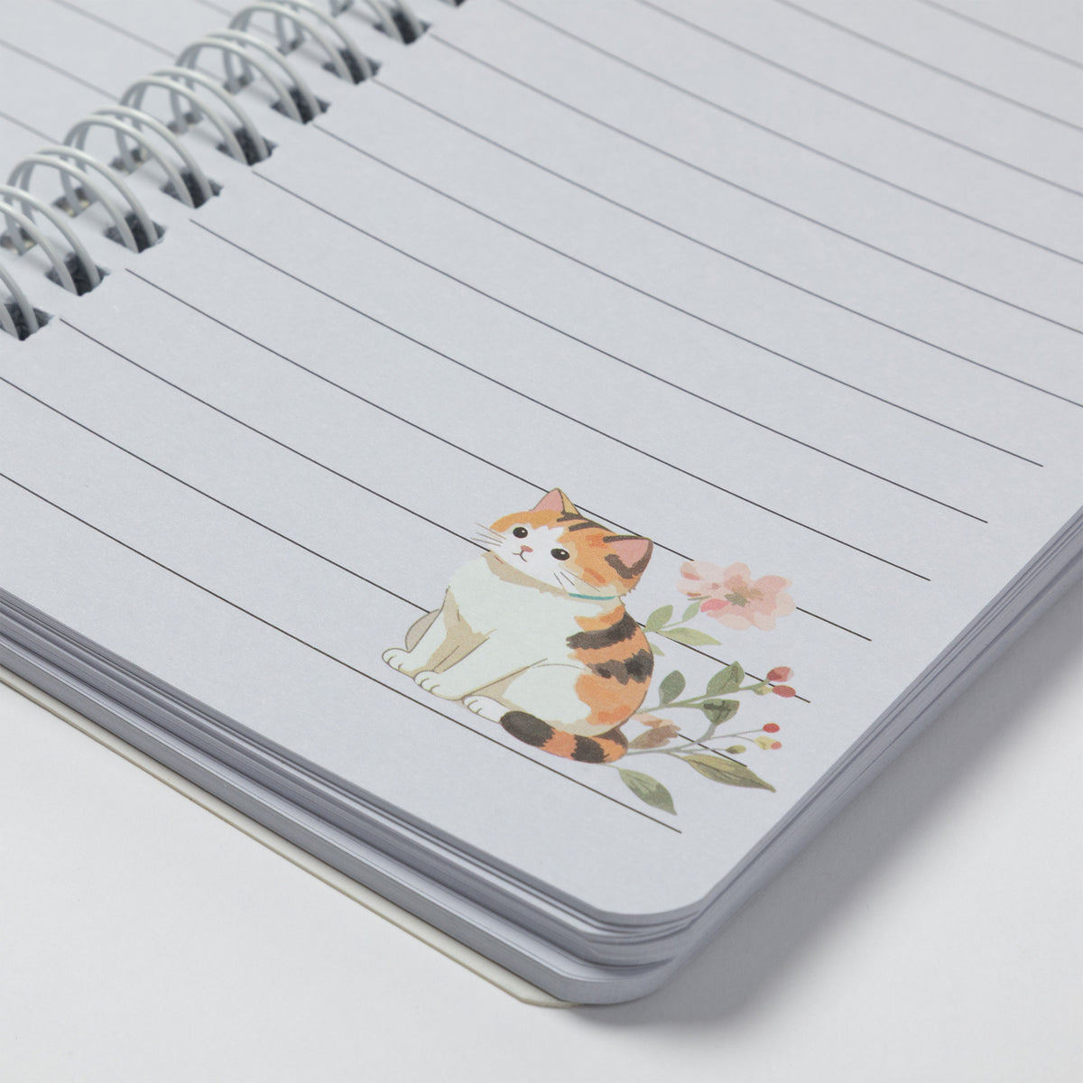 A close-up of a spiral-bound lined notebook with a small illustrated calico cat and pink flowers printed on the page.