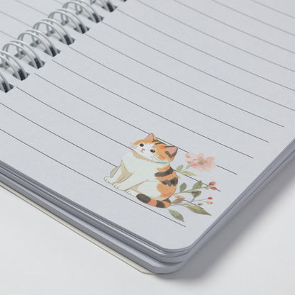 A close-up of a spiral-bound lined notebook with a small illustrated calico cat and pink flowers printed on the page.