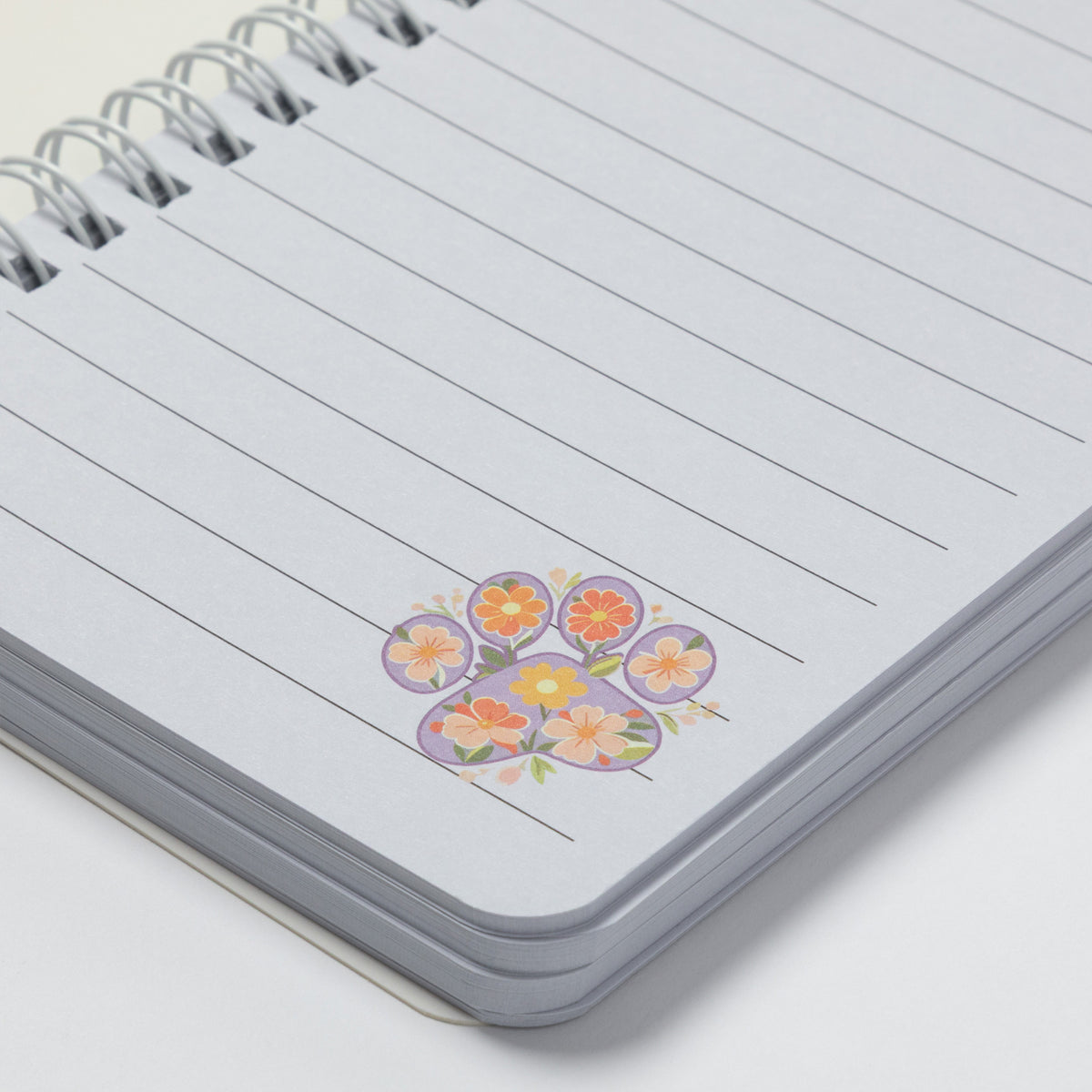 A close-up of a spiral-bound lined notebook with a small colorful floral sticker or illustration near the bottom right of the page, photographed on a clean white background.