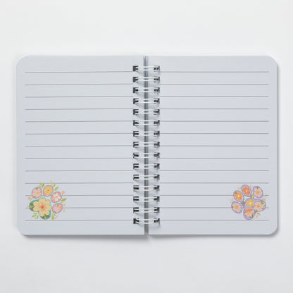 A lined notebook with a silver spiral binding, featuring small pastel floral illustrations in the bottom corners, shown open on a light gray background.