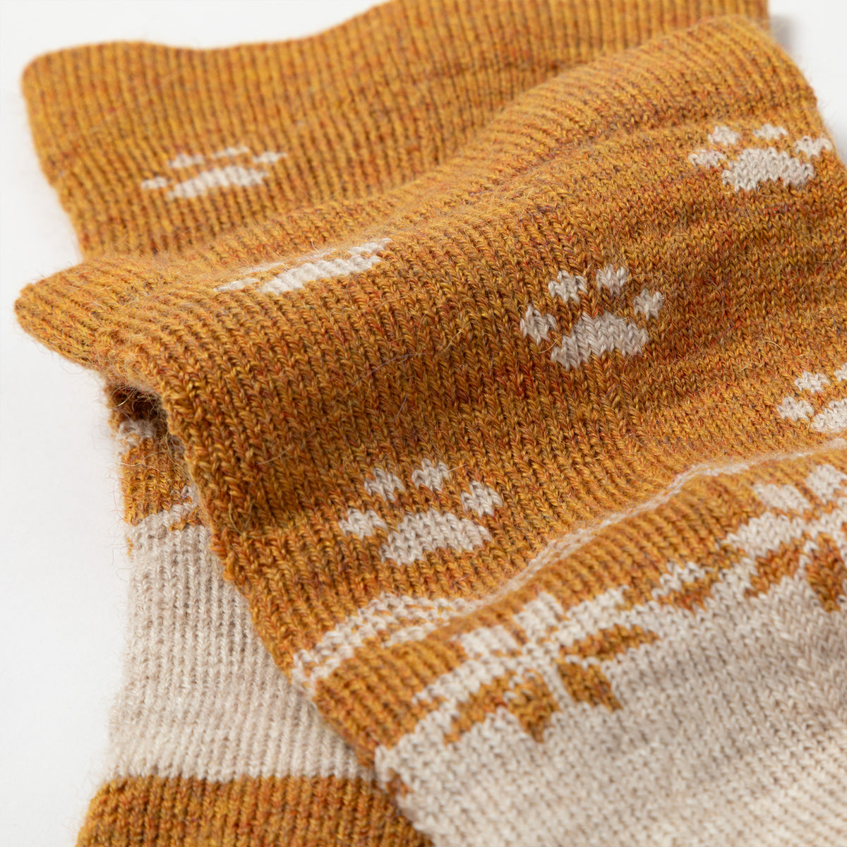 Close-up of a knit sock in a warm mustard-brown color with white paw print and snowflake patterns, folded to show the ribbed cuff and knit texture.