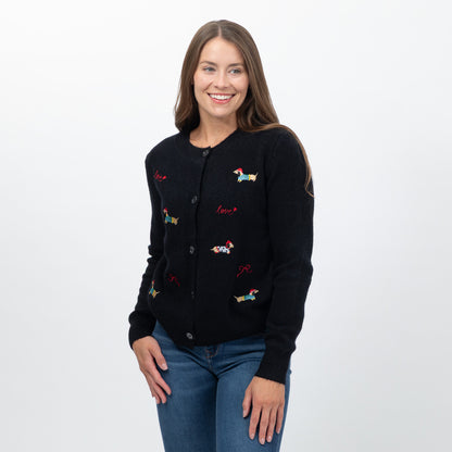 A woman wearing a black knit cardigan with colorful embroidered dog motifs and red script text, paired with blue jeans, photographed against a plain light gray studio background.