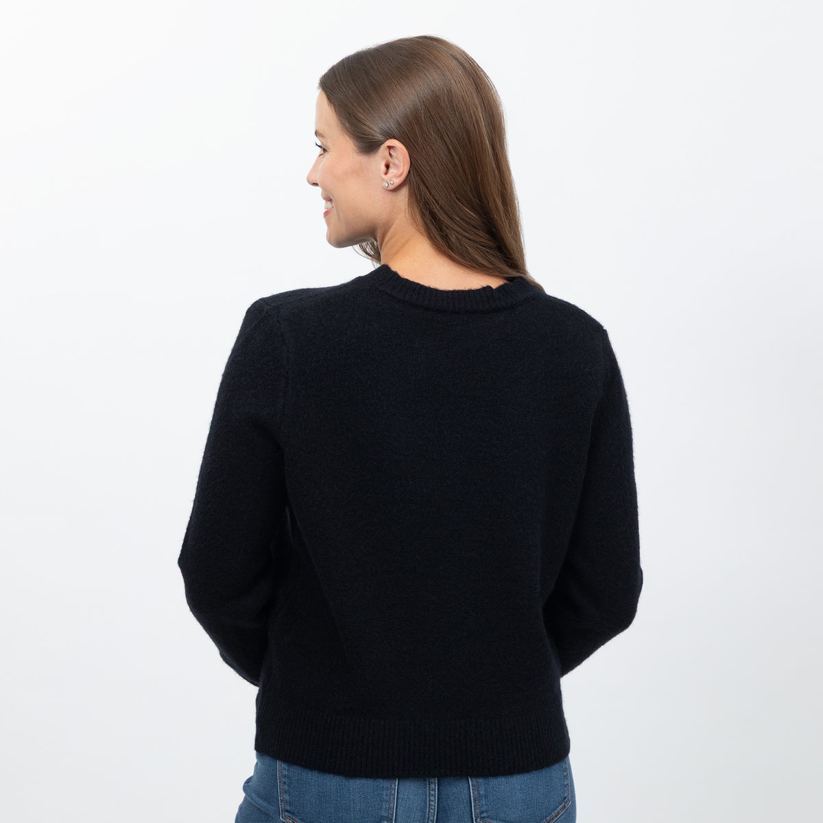 A woman viewed from the back wearing a black knit crewneck sweater and blue jeans against a plain white studio background.