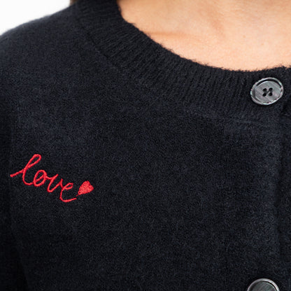 Close-up of a black knit cardigan sweater with red embroidered script reading 'love' and a small red heart on the chest, plus dark buttons.