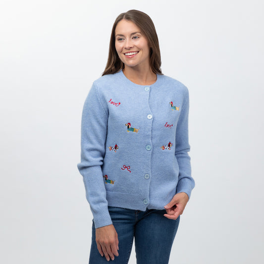 A woman wearing a light blue knit cardigan with colorful embroidered dog motifs and the word 'Love' embroidered in red, paired with dark blue jeans, against a plain white studio background.