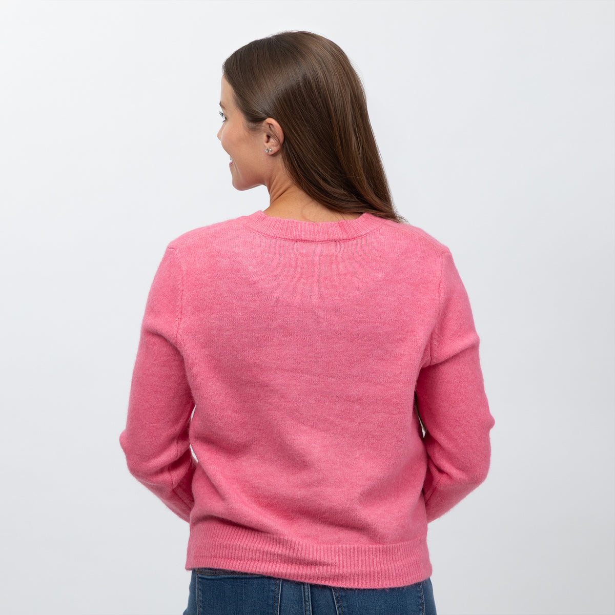 A woman viewed from the back wearing a pink knit sweater and blue jeans against a plain light gray studio background.