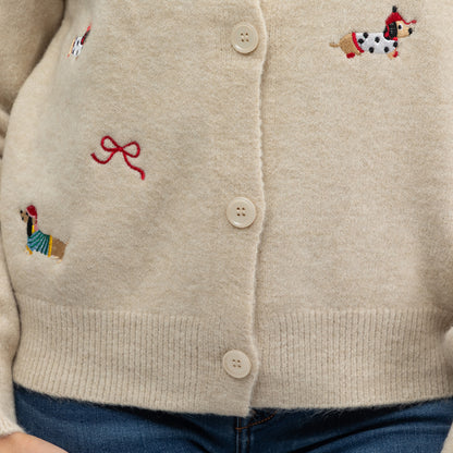 Close-up of a cream-colored knit cardigan sweater with embroidered dogs and a red bow motif, paired with blue jeans.
