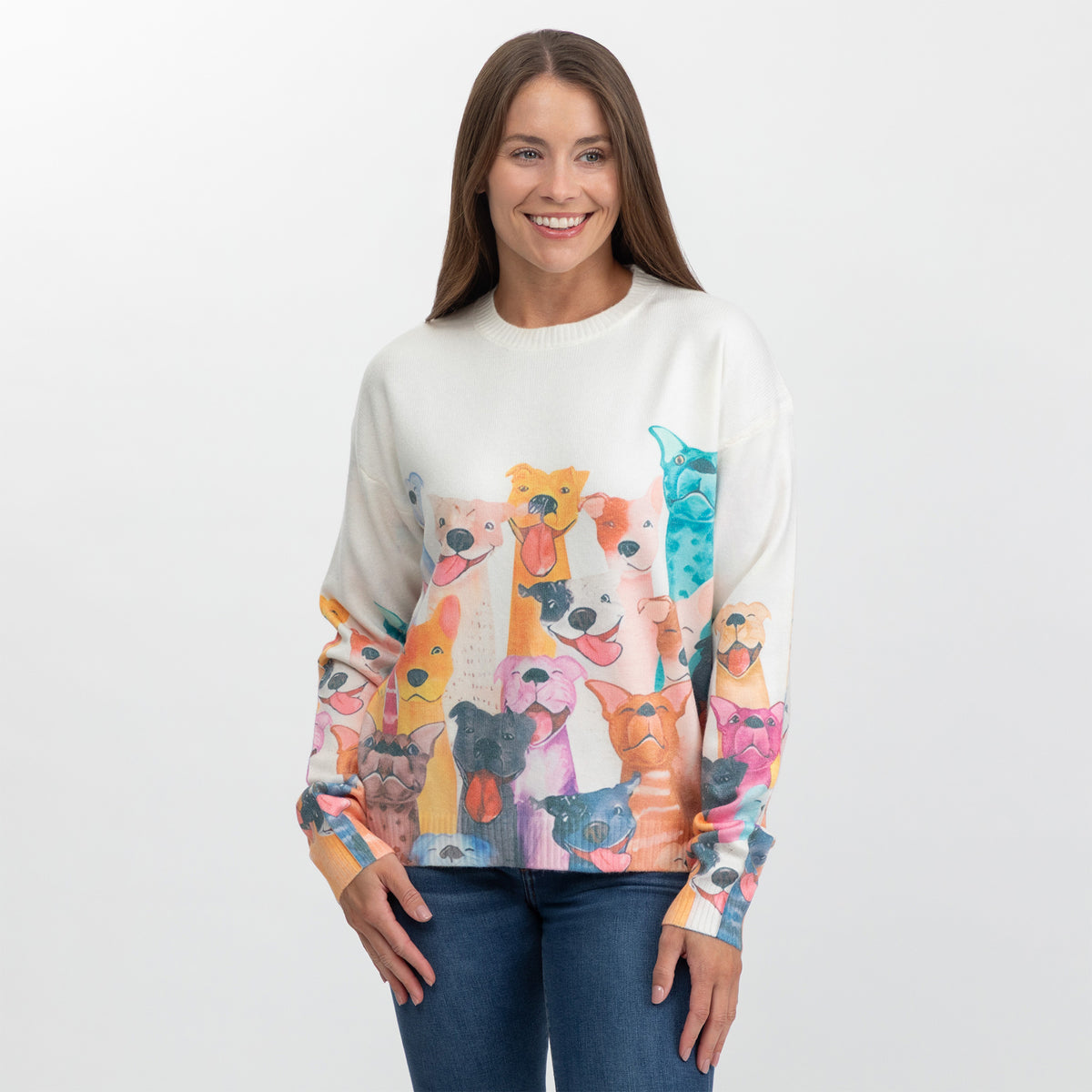 A woman wearing a cream-colored crewneck sweater featuring a colorful, all-over print of various dog faces on the front and sleeves, paired with blue jeans against a plain white studio background.