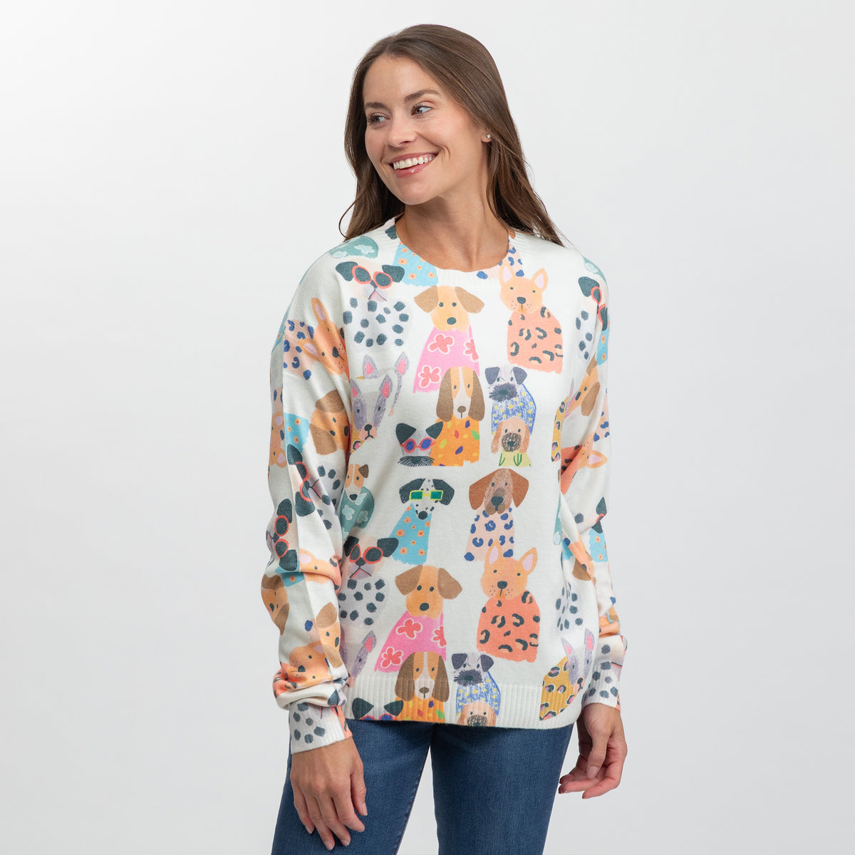 A woman wearing a cream-colored crewneck sweater with a colorful cartoon dog pattern, paired with blue jeans, photographed against a light gray studio background.