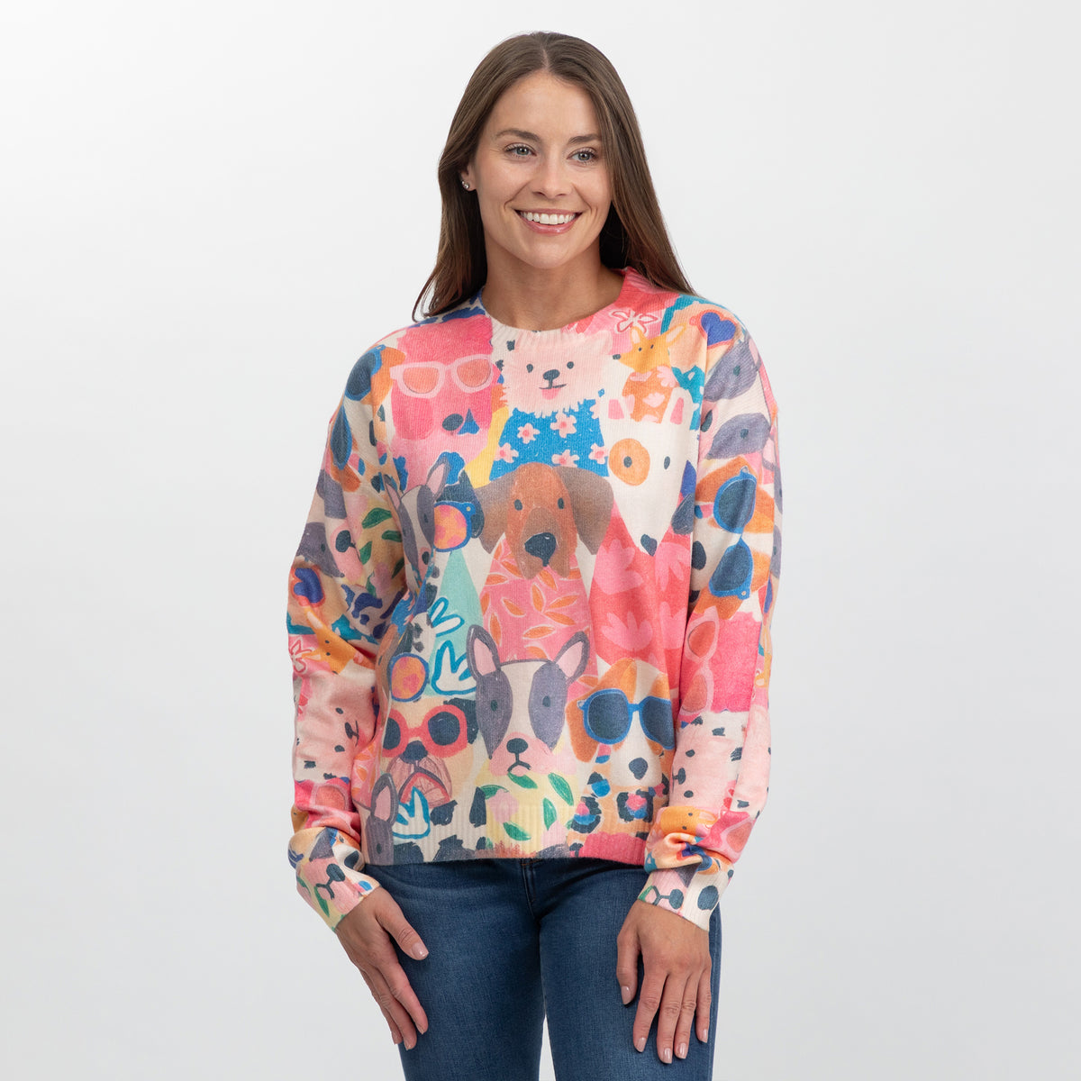 A woman wearing a long-sleeve crewneck sweater with a colorful, playful dog-themed pattern, paired with blue jeans against a plain light gray background.