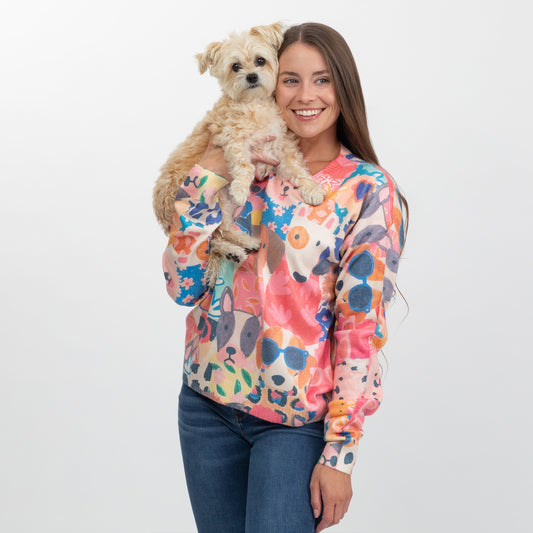 A woman holding a small fluffy dog, wearing a colorful long-sleeve crewneck sweater with a vibrant abstract floral pattern in pink, orange, blue, and yellow on a cream base, paired with blue jeans, against a plain light gray background.