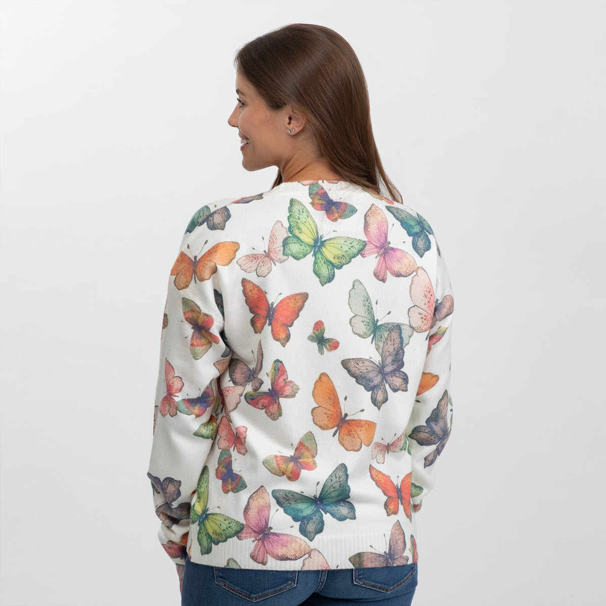 A woman shown from the back wearing a cream-colored crewneck sweater covered in a colorful watercolor butterfly print, paired with blue jeans against a plain light gray background.