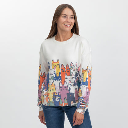 A woman wearing a white crewneck sweater with a colorful cartoon cat pattern on the lower half, paired with blue jeans, photographed against a light gray studio background.