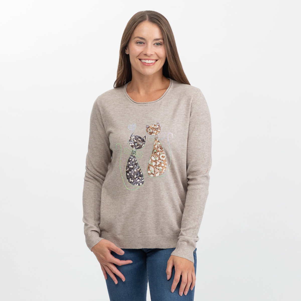 A woman modeling a light taupe crewneck sweater with a graphic of two patterned cats on the front, paired with blue jeans, against a plain white background.