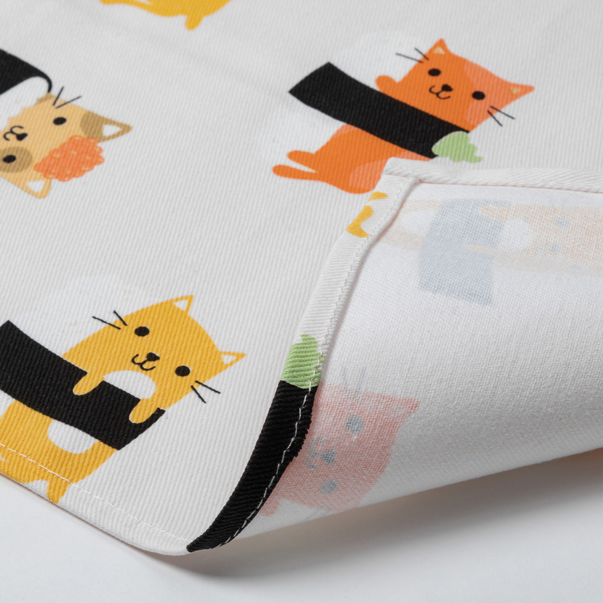 A close-up of a white dish towel, featuring a playful print of cartoon cats wrapped in black sushi rolls. The fabric is folded to show the texture and the reverse side with a subtle pastel pattern.