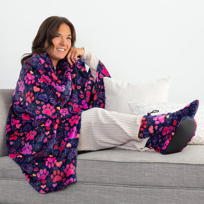 A smiling woman reclines on a gray sofa wrapped in a plush blanket and wearing matching slipper socks. The blanket and slippers feature a colorful pattern of pink hearts, purple paw prints, and orange accents on a dark navy background.