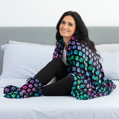 A woman sits on a bed wrapped in a black blanket featuring a colorful paw print pattern in purple, teal, and pink. She is wearing black tights and matching patterned socks.