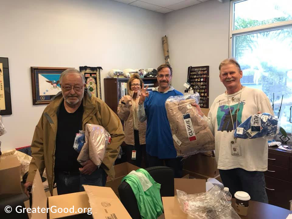 Four people holding bags of food in an office setting with GreaterGood.org branding.