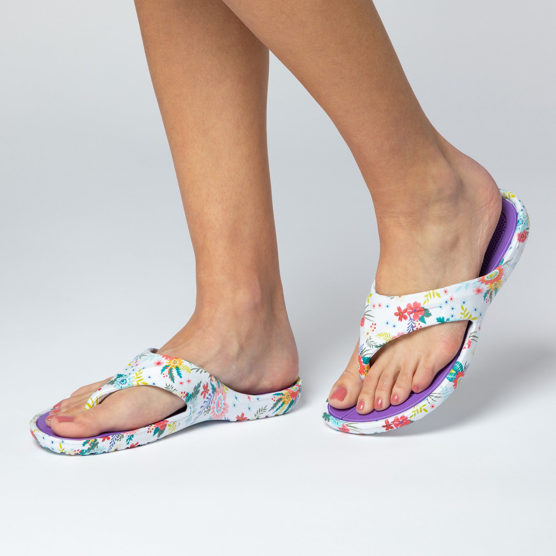 A pair of garden of paws print design EVA flip flops on a person's feet standing indoors on a white background, the garden of paws print is on a white background, and the textured foot bed is  purple