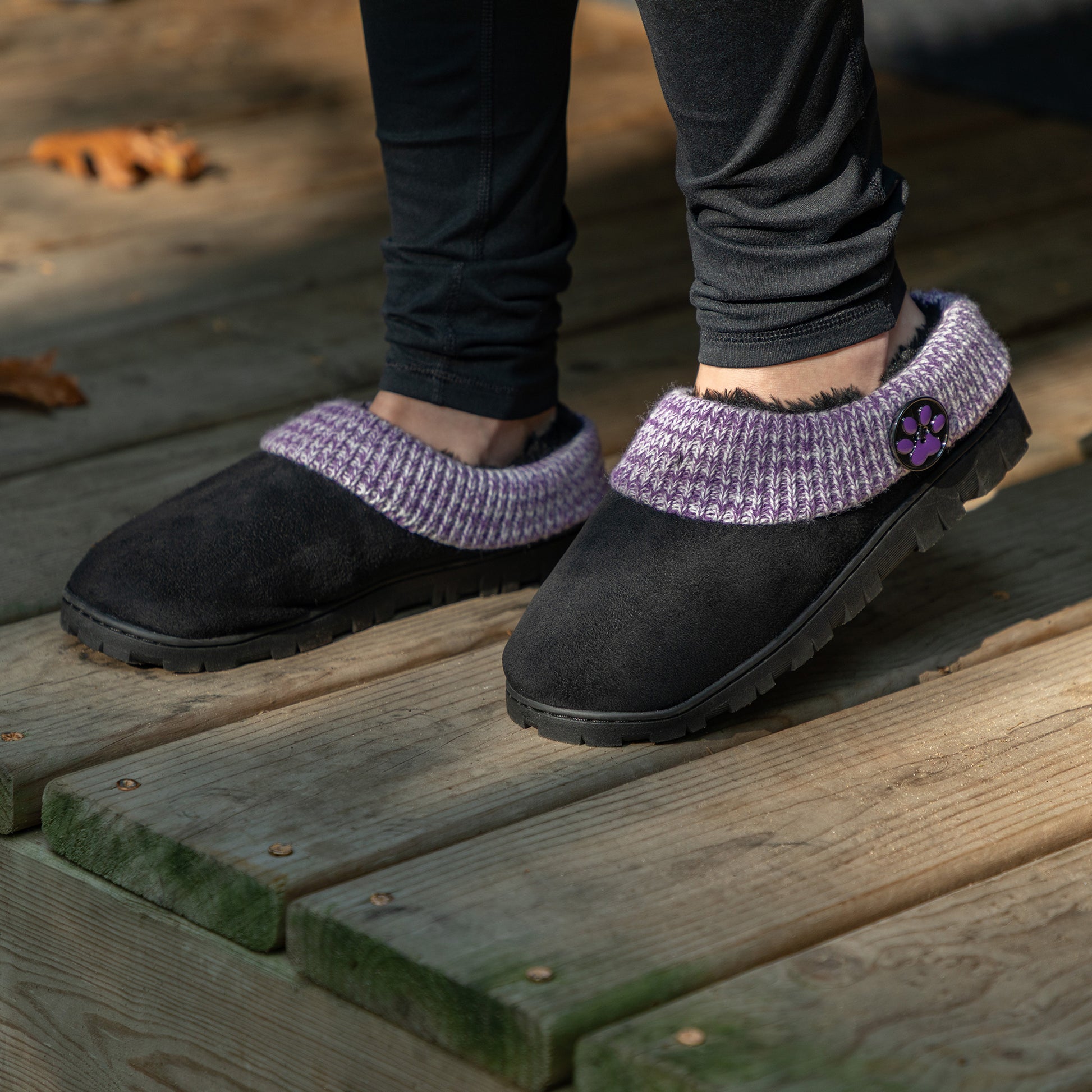 Black slippers with purple accents and black faux fur lining, on a wooden deck outdoors.