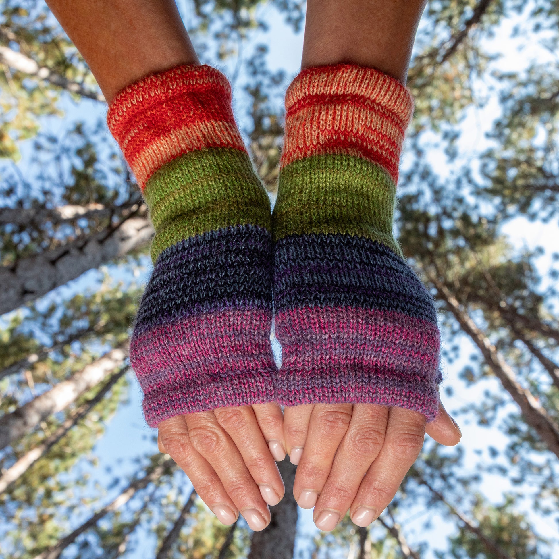 Colorful striped rainbow fingerless gloves held up against a natural background with trees. Made from alpaca wool
