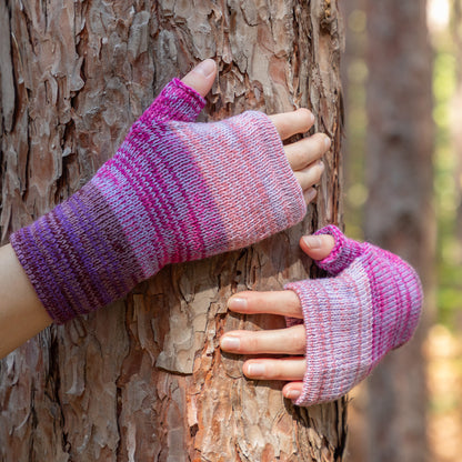 Purple and pink striped fingerless gloves on hands holding a tree trunk in a forest setting