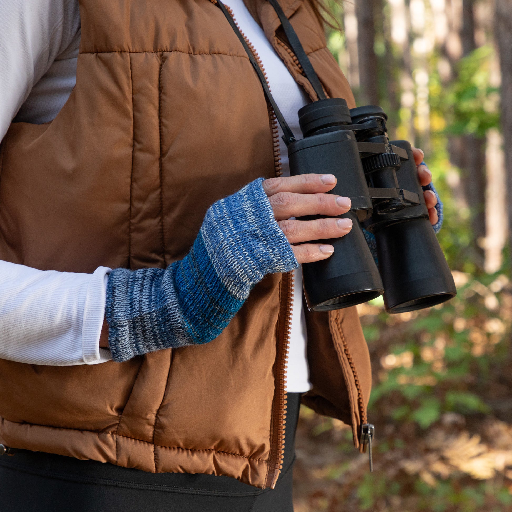 Person holding binoculars with fingerless mittens on and a forest background