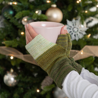 Person wearing green fingerless gloves holding a pink mug in front of a decorated Christmas tree.