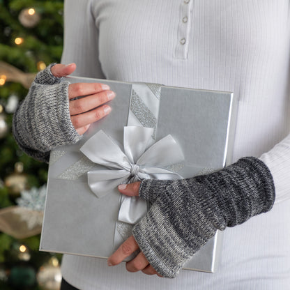Person holding a silver gift box with a bow, wearing gray fingerless gloves, against a blurred Christmas tree background.