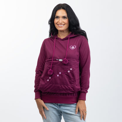 A woman wearing a maroon pullover hoodie with a front kangaroo pocket, pom-pom drawstrings, and a small embroidered heart logo on the left chest, paired with light-wash jeans against a plain white background.