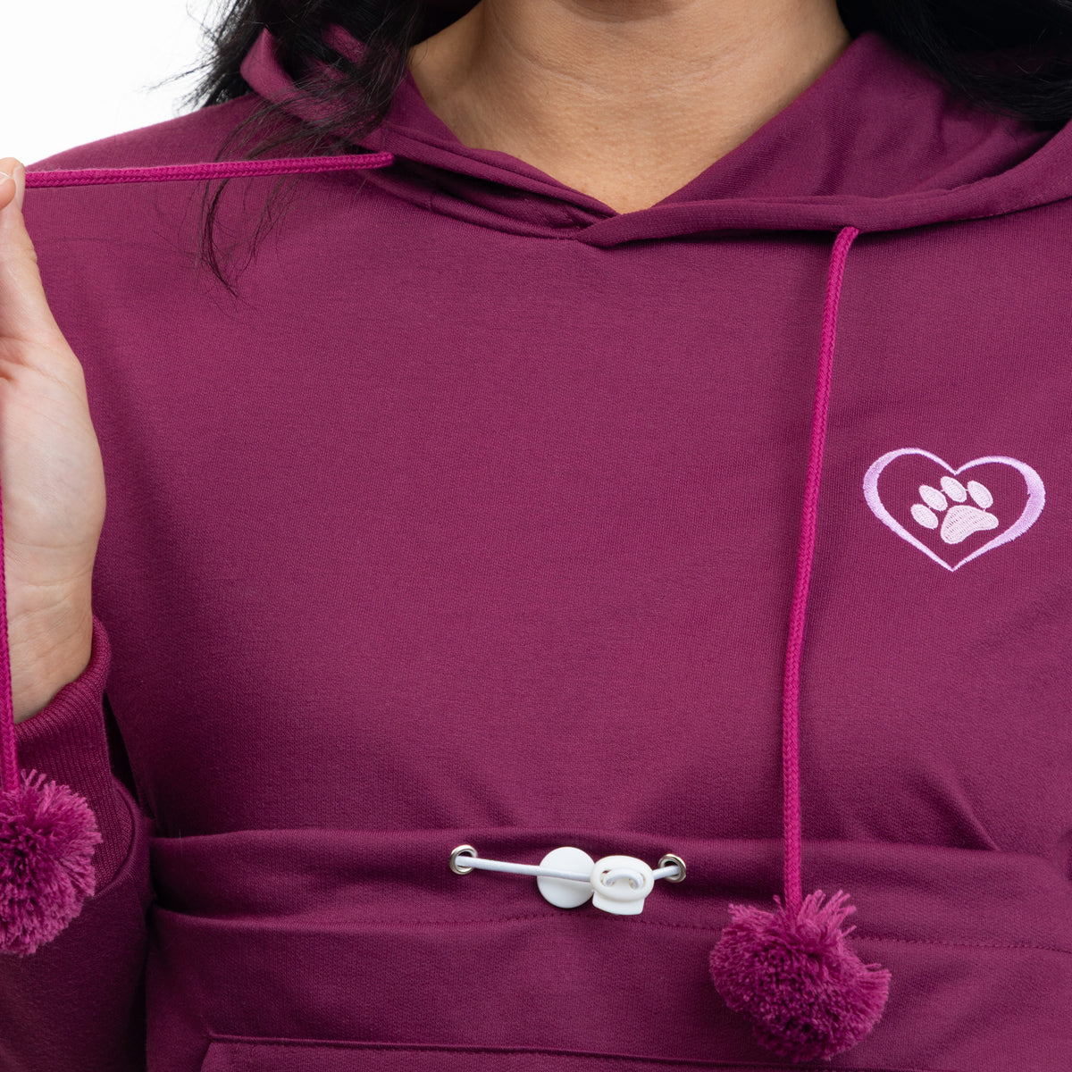 A close-up of a person wearing a deep plum purple hoodie with a white embroidered heart and paw print on the chest. The hoodie features pink drawstrings with pom-poms and a white toggle closure on the front pocket.