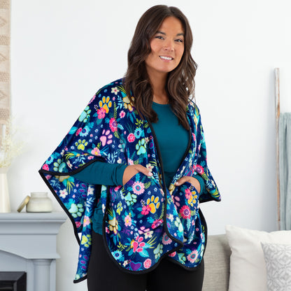 Woman in a cozy setting wearing a blue fleece, pocketed shawl, patterned with paws, flowers and foliage. The edges are lined with black piping, wrapped around her shoulders. Her hands are in both pockets.