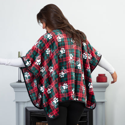 Woman in a cozy setting wearing a fleece, pocketed shawl, patterned with red and green plaid overlayed with white paws and mistletoe and black piping, wrapped around her shoulders. She is turned away to show the continuation of the pattern on the back. 